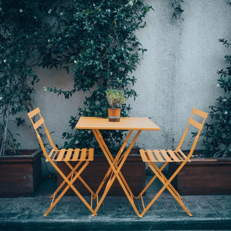 Charming outdoor bistro set with orange chairs and table, framed by lush greenery.