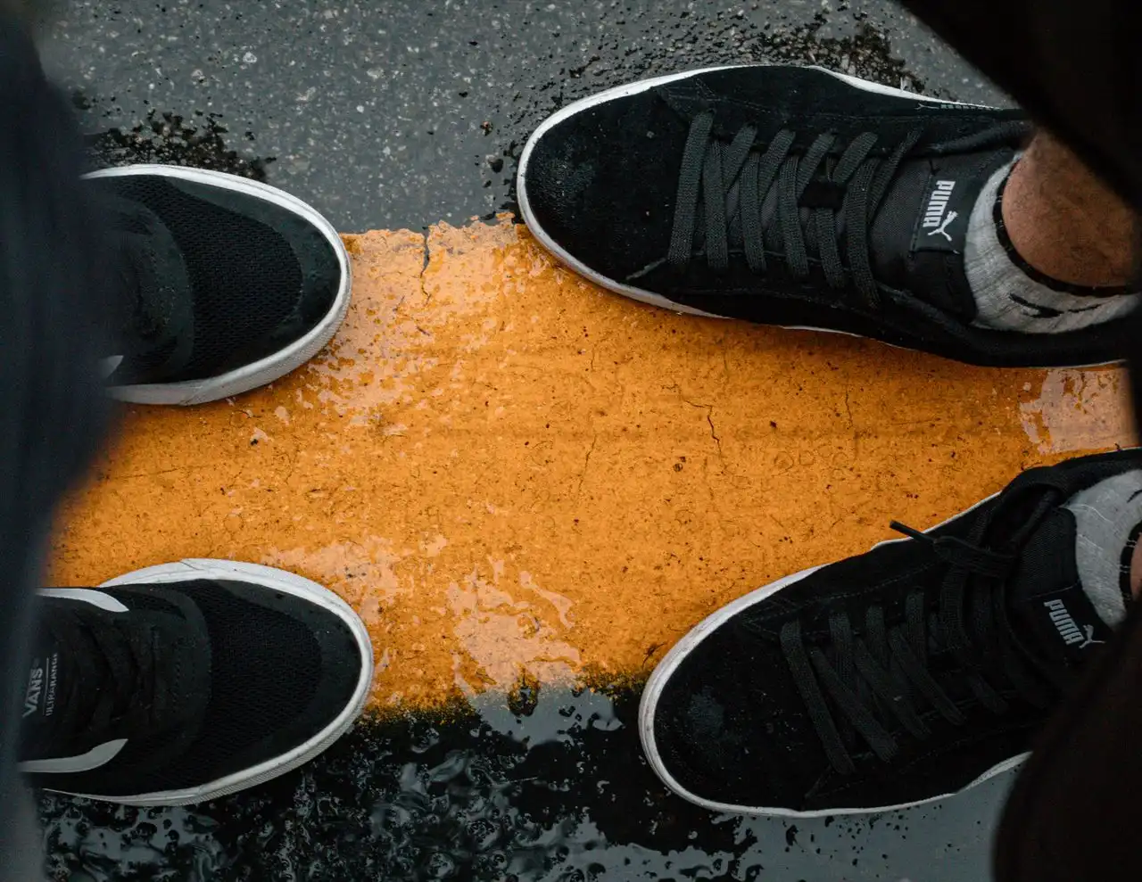 Close-up of two people wearing sneakers standing face to face on an orange road line in the rain, with wet pavement.