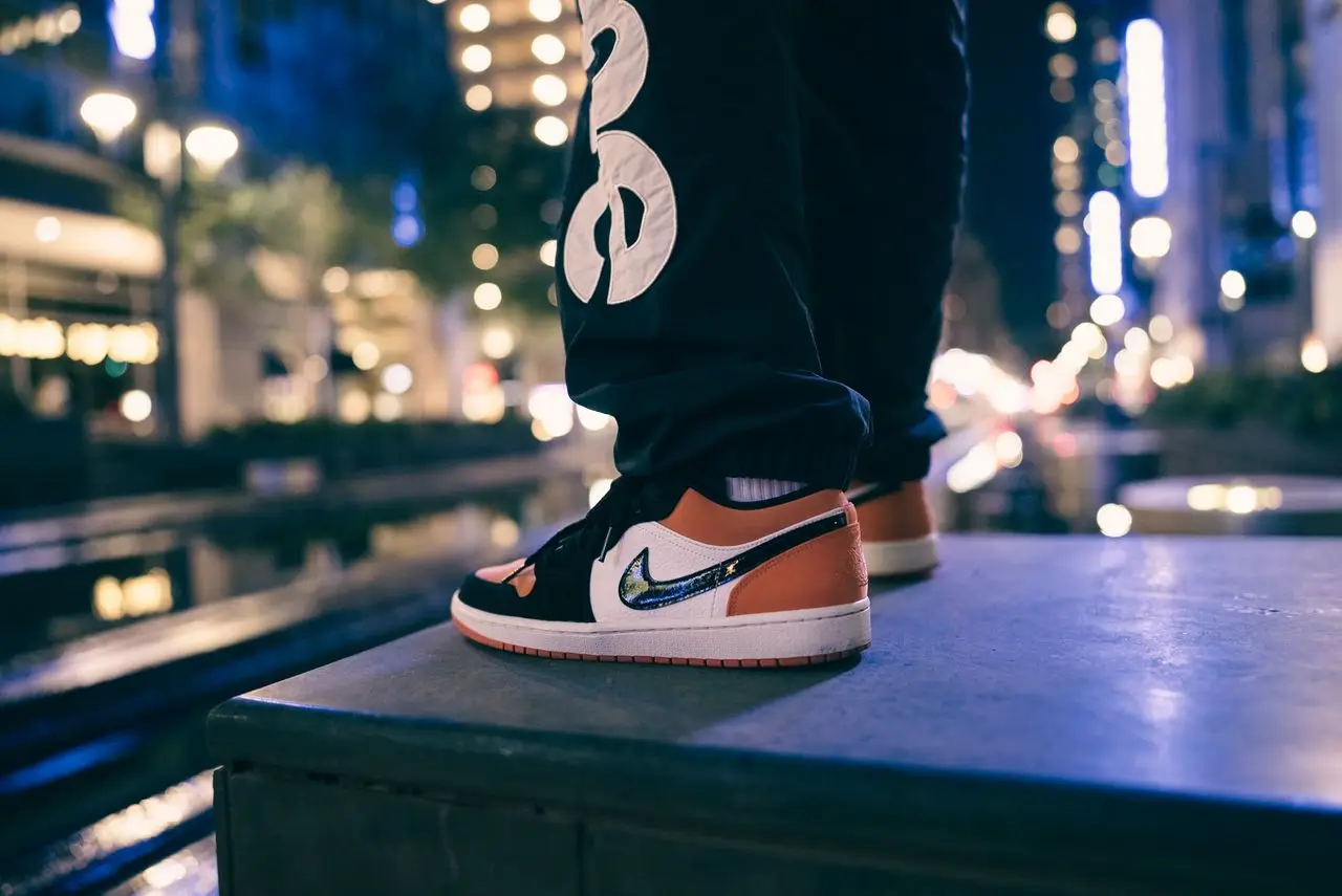 Close-up of a person wearing sneakers standing on a sidewalk wall at night with city lights and tall buildings in the background.